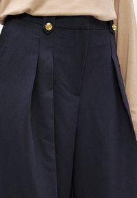 Sötétkék, magasderekú nadrág elején redőkkel, az arany spirál gombok díszítik a derékszíjat. Az anyag sima tapintásúnak tűnik.