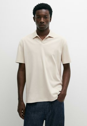 Junger Mann mit dunkler Haut und kurzen lockigen Haaren, der ein beige kurzärmeliges Poloshirt und blaue Jeans trägt, steht vor einem schlichten Hintergrund.