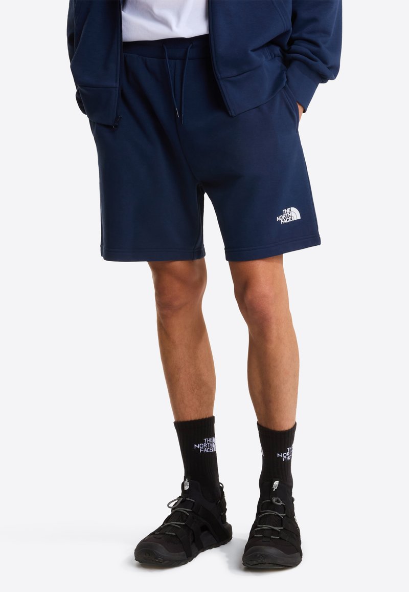 Shorts en mélange de coton bleu marine avec une ceinture élastique, des poches latérales et un logo blanc imprimé près de l'ourlet. Portés avec des chaussures et des chaussettes noires.