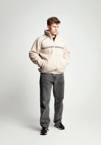 Giacca in pile beige con zip, collo alto, dettagli neri e una fascia con logo sul petto. Indossata con jeans grigi e sneaker nere.