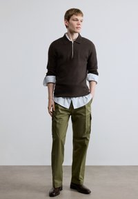 Brūns pulovers ar rāvējslēdzēju pār muguras pleciem pārvilkts pār zilru joslu kreklu, savienots ar olīvu zaļām darba biksēm. Apģērbu noslēdz brūnas kurpes.