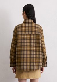 Vrouw met lang zwart haar, gekleed in een bruine en beige geruite jas en een beige rok, staande met haar gezicht naar een witte muur.