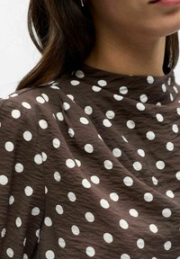 Brunt stoff med store hvite prikkete mønster på et plagg med høy hals som bæres av en person med langt mørkt hår.