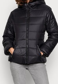 Černá prošívaná bunda s kapucí, má prošívanou texturu, přední zip a boční kapsy. Vyrobena z lesklého, hladkého materiálu.