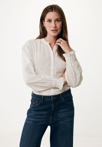 Witte blouse met lange mouwen en bloemborduursel, met knoopsluiting aan de voorkant en gerimpelde manchet, gecombineerd met donkerblauwe spijkerbroek.