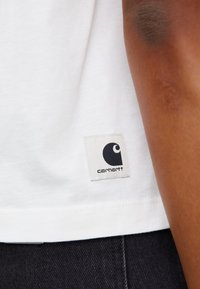 Bílý bavlněný tričko s čtvercovou nášivkou Carhartt a černým logem. Viditelné švy na lemu, nošeno s tmavými džíny.