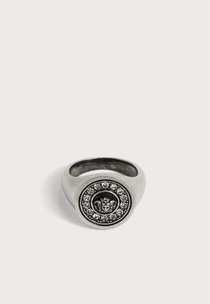 Bague en argent avec un visage circulaire présentant une tête de lion en relief entourée d’un cercle de pierres précieuses transparentes.