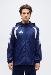 Νεαρός άνδρας που φοράει ένα σκούρο μπλε αδιάβροχο μπουφάν Adidas με λευκές ρίγες, στέκεται μπροστά σε ένα απλό φωτεινό φόντο, στραμμένος προς τα εμπρός.