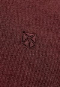 Gros plan sur un tissu tricot bordeaux avec un petit logo géométrique en forme de bouclier brodé dans une couleur de fil assortie.