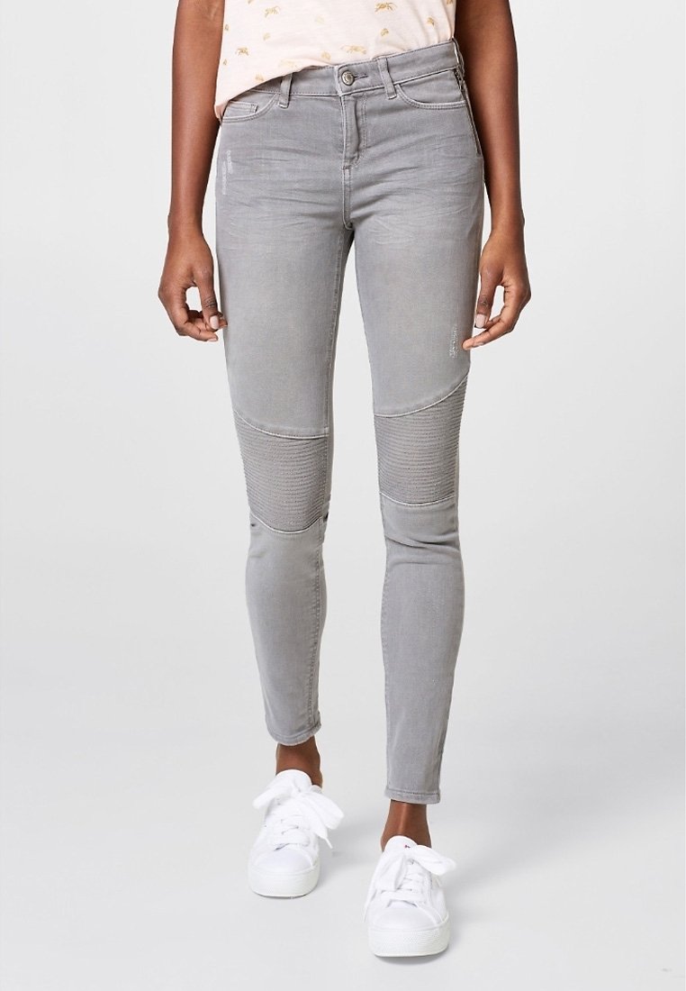 Jean skinny gris avec des détails de panneau au niveau des genoux, présentant une texture lisse et un léger effet délavé. Associé à des baskets blanches pour le contraste.