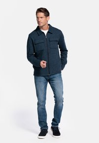 Donkerblauwe ritsjack met twee borstzakken, gedragen over een witte t-shirt, gecombineerd met lichtblauwe jeans en zwarte sneakers.