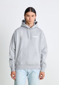 Grå hoodie med känguruficka, justerbara snoddar och vit logotyp på bröstet; kombinerad med ljusblå jeans.