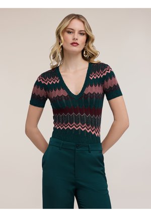 Lyshåret kvinde med løse krøller bærer guldbøjler, en V-hals kortærmet strikbluse med lyserøde og mørkegrønne zigzag-mønstre og mørkegrønne bukser.