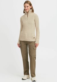 Beige Kabelstrickpullover mit hohem Kragen, Reißverschluss und gerippten Bündchen, kombiniert mit olivgrünen Cargo-Hosen und weißen Turnschuhen.