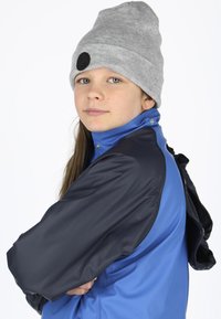 Blå og sort jakke med høj krave og trykknapper, båret med en grå strikket hue. Glat tekstur og afslappet design.