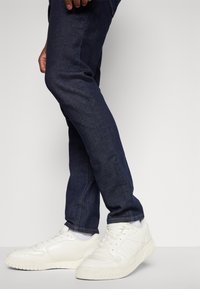 Sötétkék farmernadrág, slim fit fazonban, fehér sportcipőkkel, amelyek texturált oldalakkal és fűzővel rendelkeznek, sima háttér előtt.