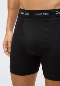 Homem vestindo boxers pretos da Calvin Klein, exibindo o logotipo na cintura e detalhes de costura texturizada na parte da frente e nas pernas.