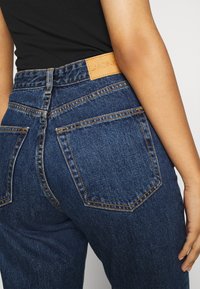 Pantalones vaqueros de mezclilla azul de tiro alto con una etiqueta de cuero marrón, costuras naranjas y dos bolsillos traseros con un diseño clásico de cinco bolsillos.