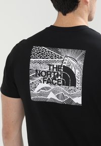 Černé tričko s bílou grafikou na zadní straně, zobrazené abstraktní krajiny a text "THE NORTH FACE" v tučných písmenech.