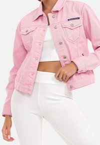 Rosa denimjacka med silverfärgade tryckknappar, två bröstfickor och en cropped design, bärs över en vit ribbad topp och byxor.