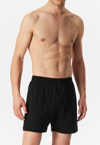 Boxer neri in cotone con un bottone e una vita elastica, mostrati su un modello maschile con uno sfondo neutro.