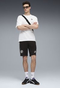 Vit bomull t-shirt, svarta shorts med sidodetaljer och logotyper, svarta sneakers och solglasögon. Modellen står med armarna i kors, neutral bakgrund.