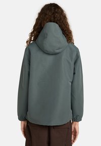 Person mit langen lockigen Haaren, die eine blau-graue Kapuzenjacke und dunkle braune Hosen trägt, zeigt mit dem Rücken zu einem schlichten Hintergrund.