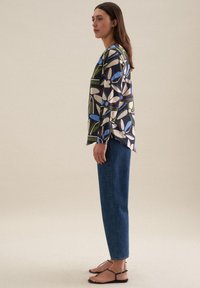 Blusa a maniche lunghe con un motivo floreale multicolore su sfondo scuro, abbinata a jeans blu a gamba dritta e sandali neri.