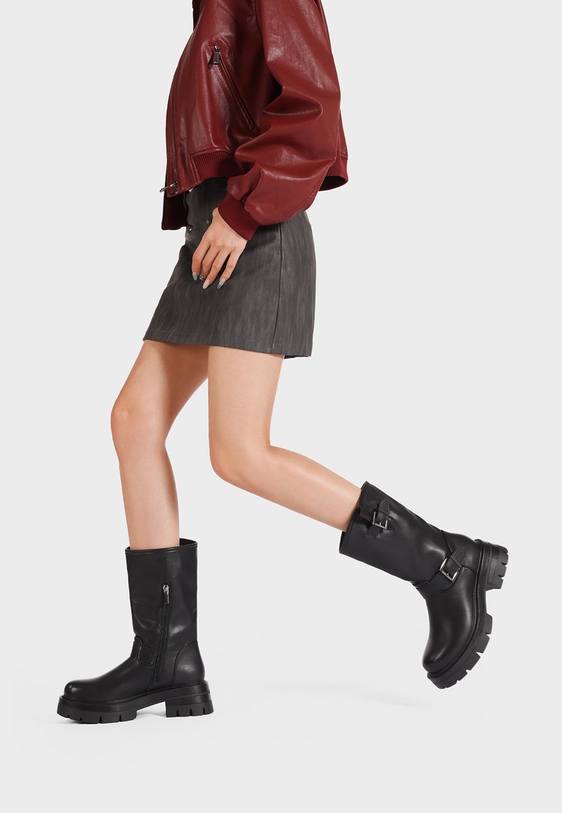 Stivali in pelle nera alla caviglia con punta arrotondata, suola in gomma spessa, zip laterale e dettaglio con fibbia. Indossati con una gonna grigia e una giacca rossa.