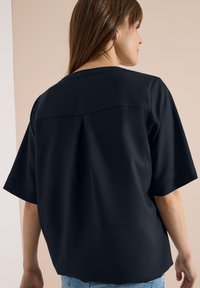 Femme aux cheveux bruns lisses, portant un t-shirt noir ample à manches courtes, vue de dos sur un fond beige.