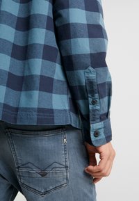 Camicia a quadri blu e navy con una texture morbida, caratterizzata da un orlo dritto e polsini con bottoni, abbinata a jeans azzurri chiari.