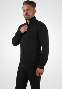 Schwarzer Reißverschluss-Pullover mit gerippter Textur, hohem Kragen und enganliegenden Bündchen, getragen über einem weißen Hemd, kombiniert mit dunklen Hosen.