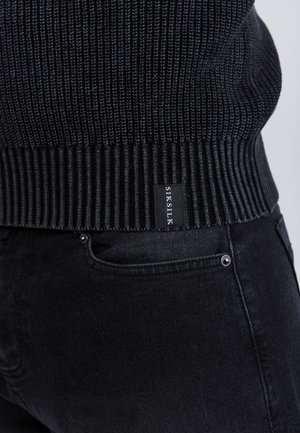 Zwarte ribgebreide trui met een tekstuur aan de onderkant en een logo-label. Gecombineerd met donkere jeans, die de gladde stofstructuur en de zakdetails benadrukken.