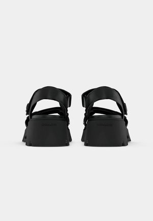 Par sorte platformsandaler med tykke hæle og justerbare bagremme, vist bagfra på en ensfarvet baggrund.