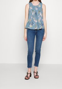 Sininen kukallinen hihaton toppi, jossa on poimutuksia ja vaaleanpunaisia sekä vihreitä kuvioita. Yhdistetty tummansinisiin kapeisiin farkkuihin ja mustiin hihna sandaaleihin.