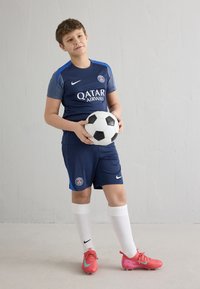 Mėlyna „Nike“ futbolo marškinėlių su logotipu, juodos trumpikės, juodai baltas futbolo kamuolys, rožinės futbolo bateliai ir balti kojinės. Neutralus fonas.