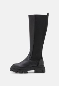 Botas negras hasta la rodilla con una parte delantera de cuero suave y una parte trasera de tela texturizada, con una suela gruesa estriada y una lengüeta para facilitar su uso.