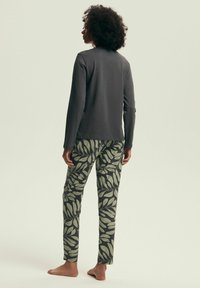 Ciemnoszara bluzka z długim rękawem w połączeniu z zielonymi spodniami w liściowy wzór. Tkanina wydaje się miękka i lekka. Modelka stoi boso, widok z tyłu.