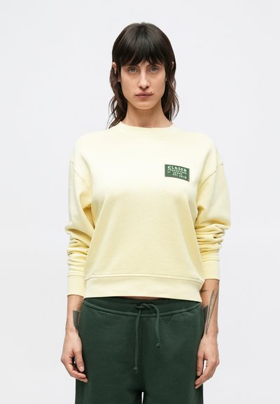 Sweat-shirt jaune à col rond, manches longues, avec un patch vert comportant du texte. Associé à un pantalon de survêtement vert foncé. Design décontracté, tissu doux.
