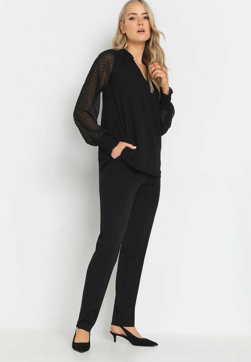 Blusa negra con mangas transparentes texturizadas, escote en V y bolsillos, combinada con pantalones negros a medida y tacones negros de punta.
