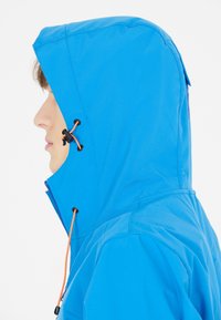 Blå vattentät jacka med huva, orange dragsko och slät textur. Sidovy kan visa design detaljer och sömmar.