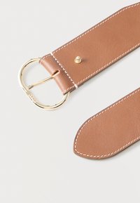 Καφέ δερμάτινη ζώνη με λευκή ραφή, χρυσή στρογγυλή αγκράφα και μεταλλικό πείρο για το κλείσιμο, σε λευκό φόντο.