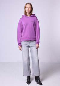 Felpa viola con coulisse, con testo sul davanti. Abbinata a jeans grigi chiari a gamba larga e stivaletti neri su uno sfondo neutro.