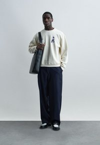 Άνδρας που φοράει κρεμ φούτερ με μικρό γραφικό, ευρύ παντελόνι σε μπλε ναυτικό με ρίγες, μαύρα και άσπρα μοκασίνια, κρατώντας μια μεγάλη μαύρη τσάντα ώμου.