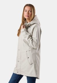 Mladá žena v světle béžovém plášti s kapucí a druky, drží límec, stojí před jednobarevným pozadím.