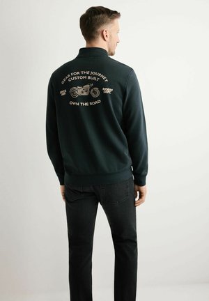 Mężczyzna w ciemnej kurtce z grafiką motocykla i napisem "Wyposażenie na podróż, wykonane na zamówienie, zdobywaj drogę" na plecach, zwrócony tyłem.