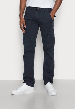 Hombre con pantalones cargo azul marino oscuro con bolsillos laterales con solapa y zapatillas blancas, de pie con las manos en los bolsillos contra un fondo liso.