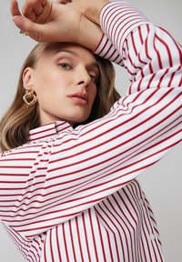 Kobieta nosząca złote kolczyki w kształcie serca oraz białą koszulę w czerwone paski, pozująca z uniesionymi rękami na jednolitym tle.