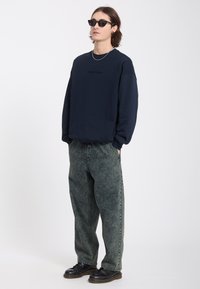 Homme portant des lunettes de soleil noires, un sweat-shirt bleu marine, un jean délavé à l'acide, des chaussures noires et un collier chaîne en argent, debout les mains dans les poches.