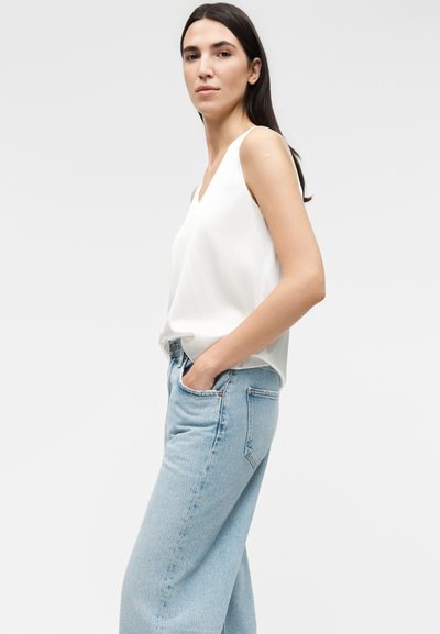 Γυναίκα με μακριά σκουρόχρωμα μαλλιά, που φοράει λευκό αμάνικο μπλουζάκι και ανοιχτά μπλε τζιν με ψηλή μέση, με τα χέρια στις τσέπες.